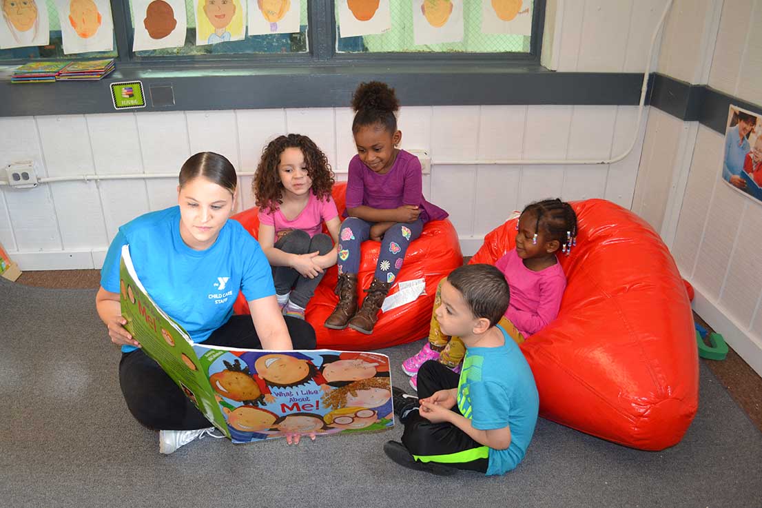 A childcare teacher is sitting on the floor of a classroom holding a book. Young children are gathered around looking at the book